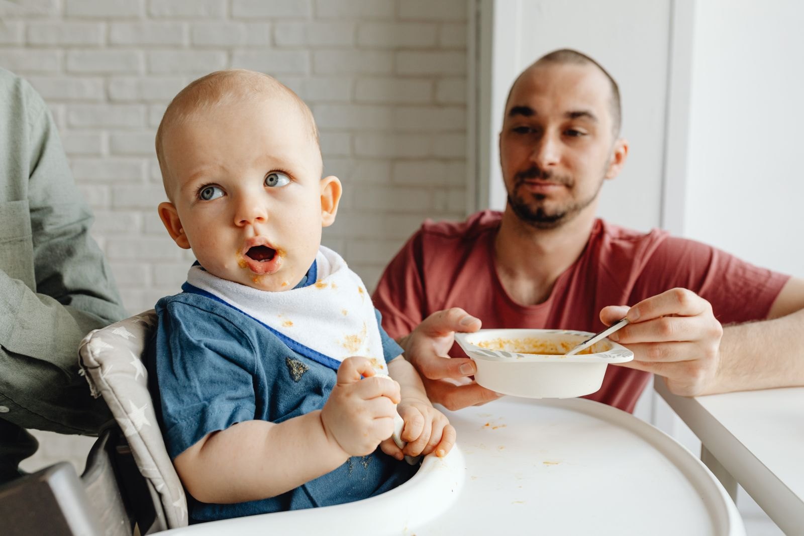 Ein Vater füttert sein Baby im Hochstuhl mit einem Löffel Brei
