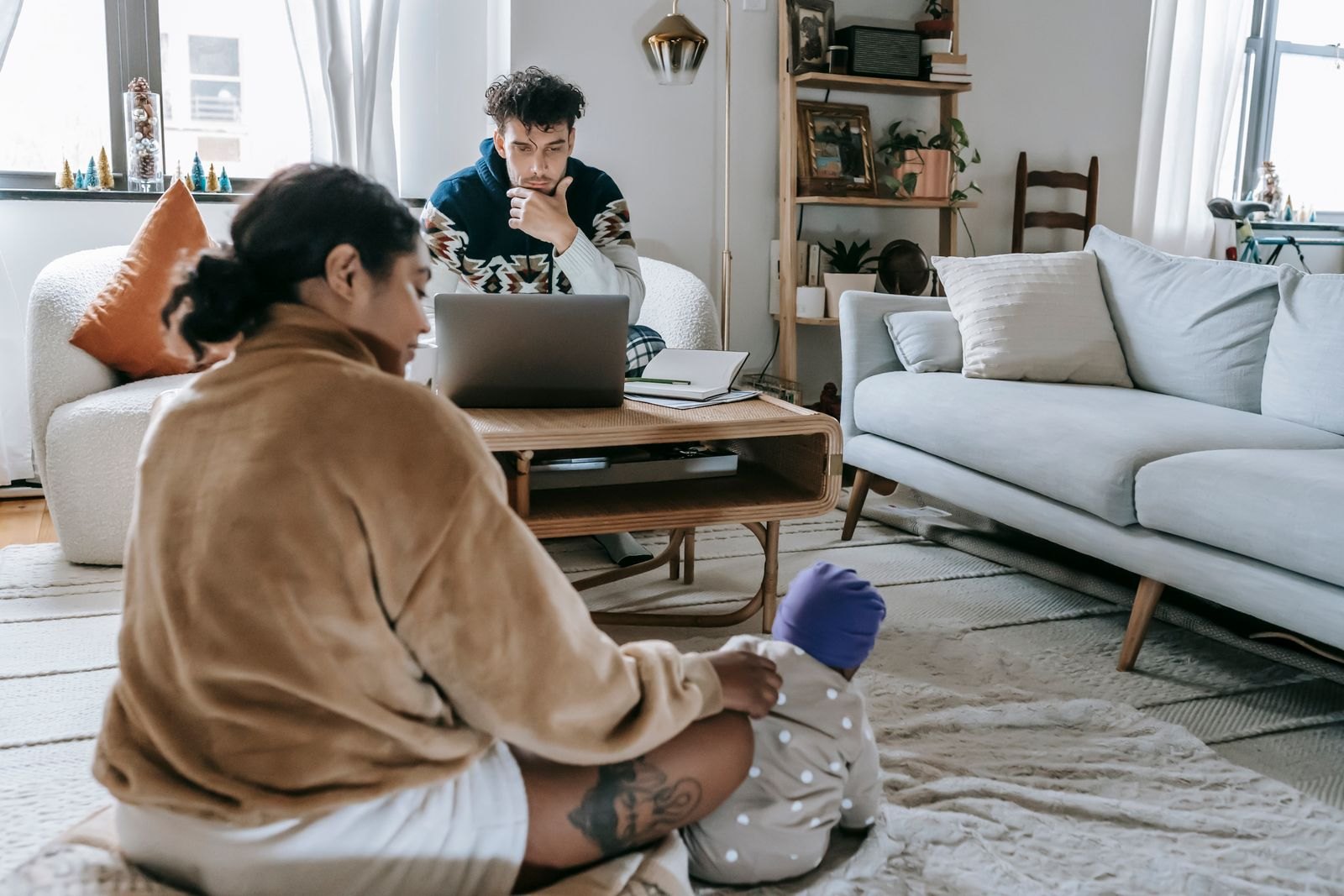 Die Mutter kümmert sich auf dem Boden ums Baby, während der Vater im Hintergrund am Laptop sitzt