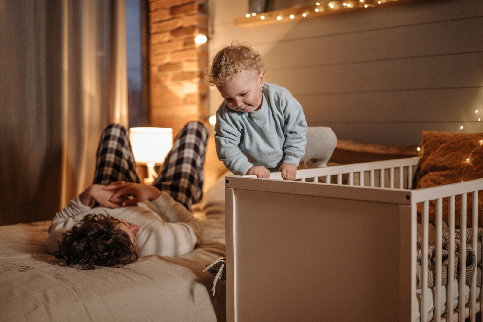Ein Elternteil liegt abends im Bett, während ein Kleinkind sich am Gitterbett hochzieht
