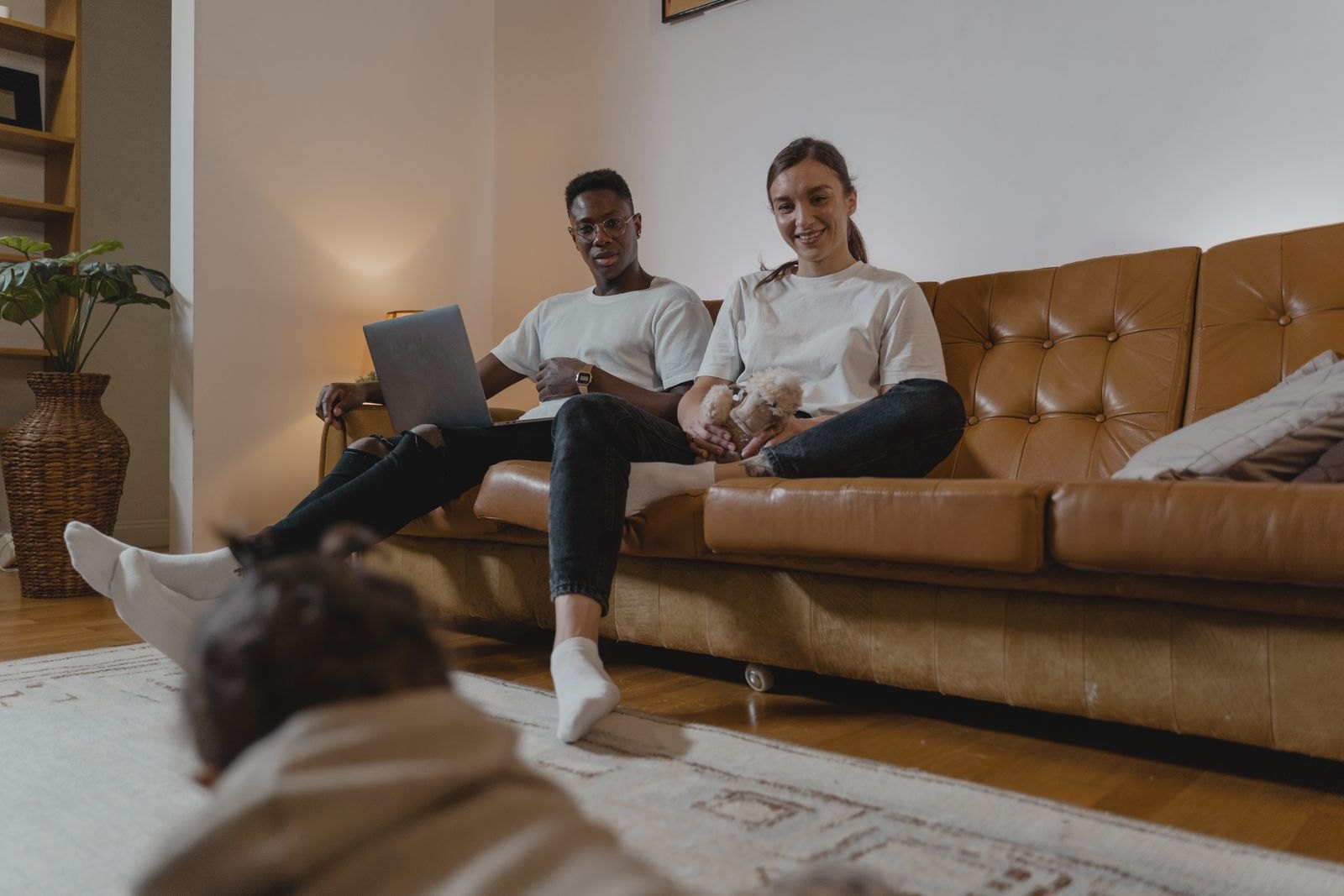 A couple sitting together on the couch while their baby explores nearby on the floor