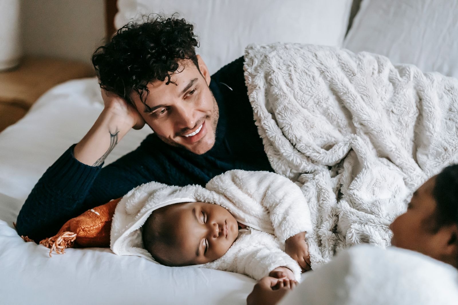 A father lying with the baby while his partner rests nearby in the background
