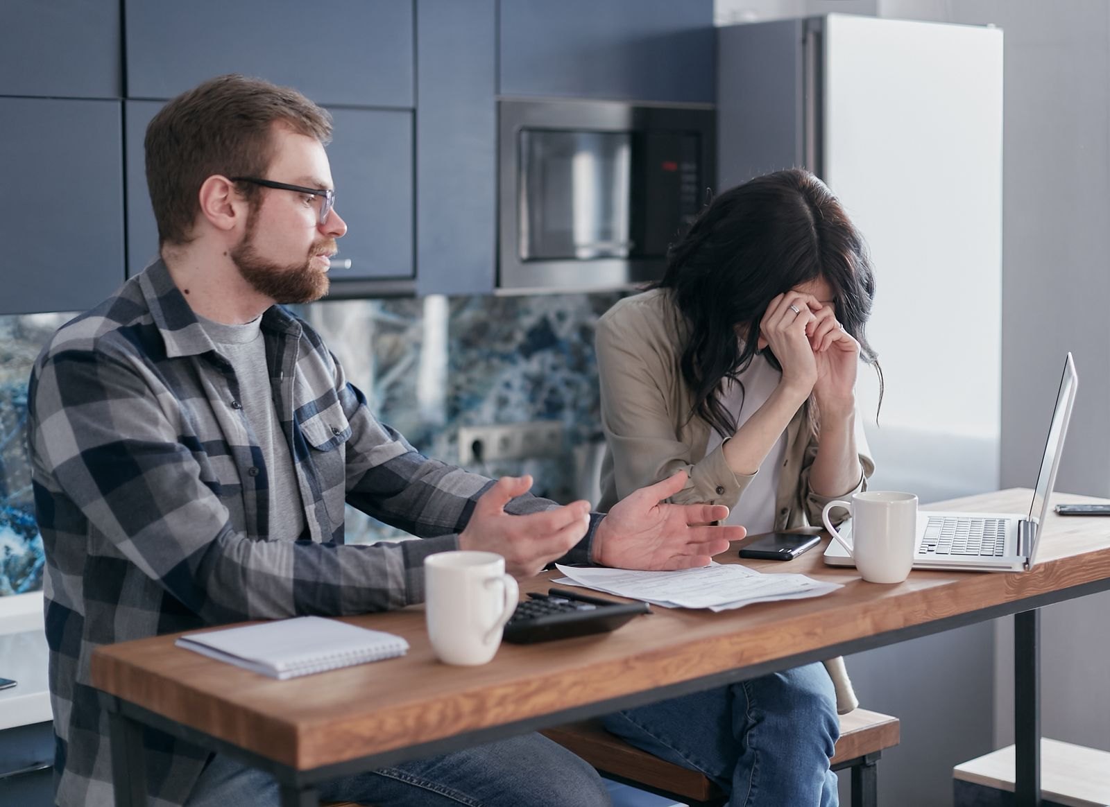 A man sipping coffee with a skeptical expression while his partner hands him household paperwork across the table