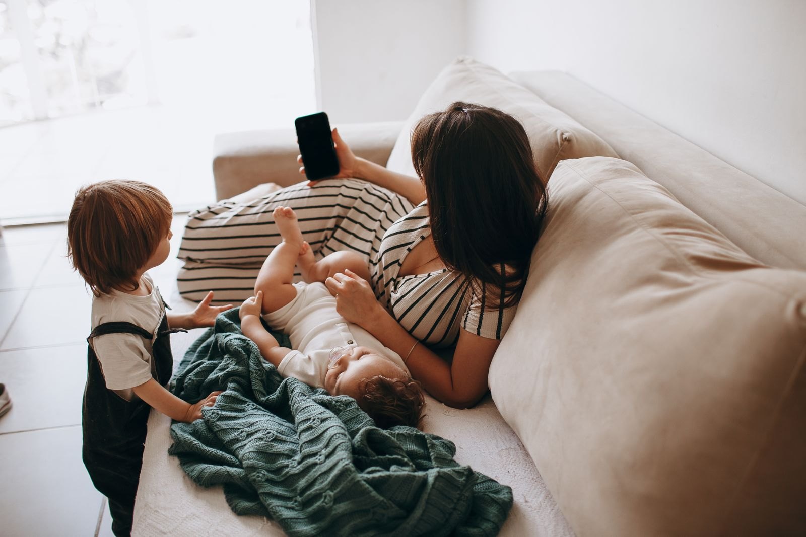 A tired parent lying on a couch with a baby, phone in hand, a toddler nearby