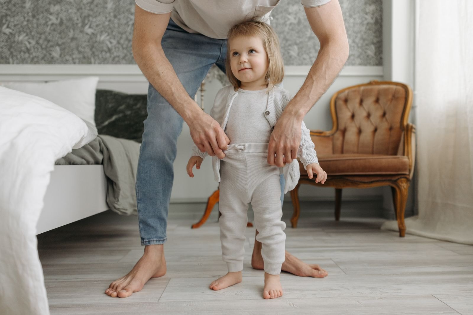 A parent helping a toddler get dressed while the toddler stands and looks up