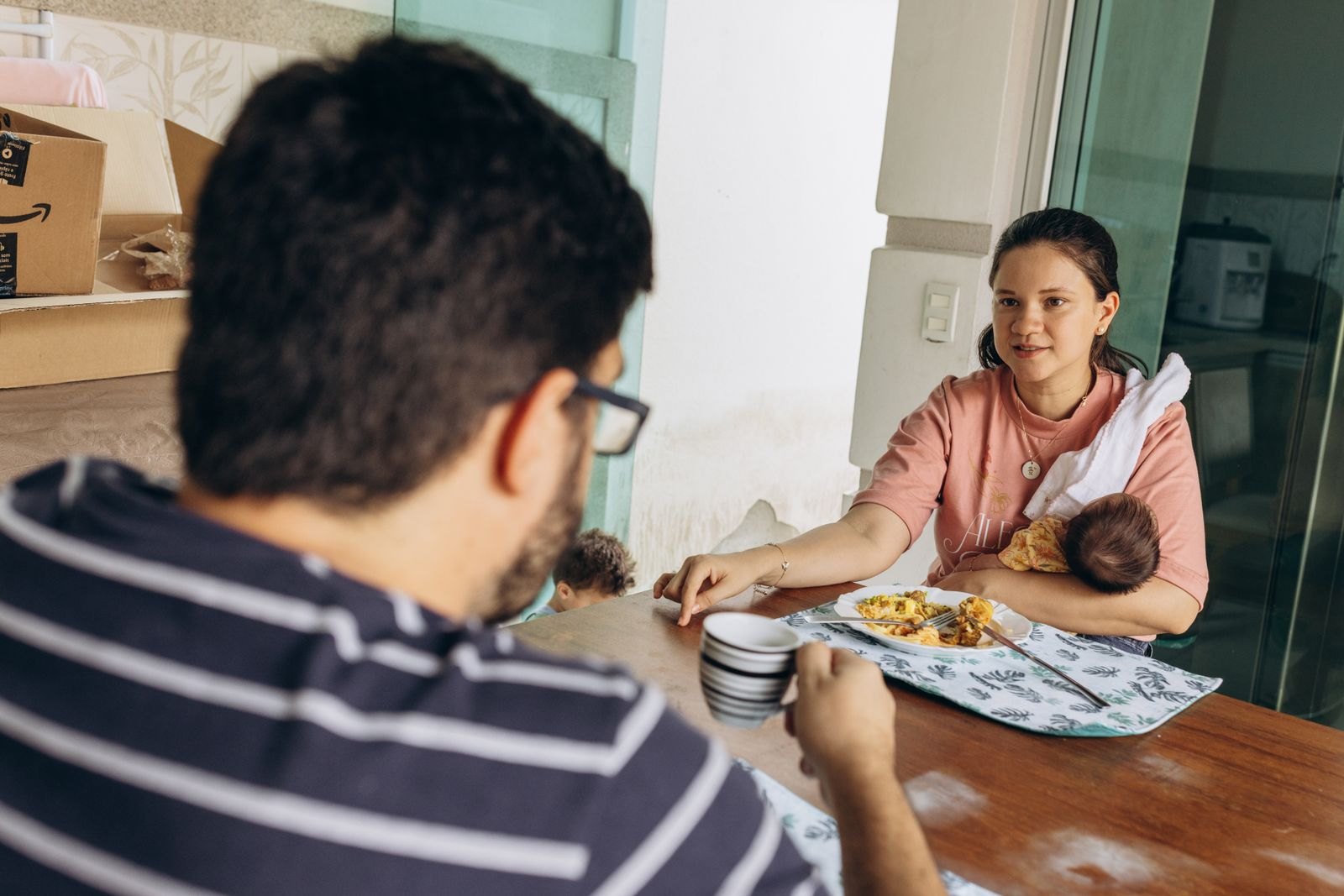 Un padre toma café en primer plano mientras, al otro lado de la mesa, la madre sostiene al bebé recién nacido y habla con él