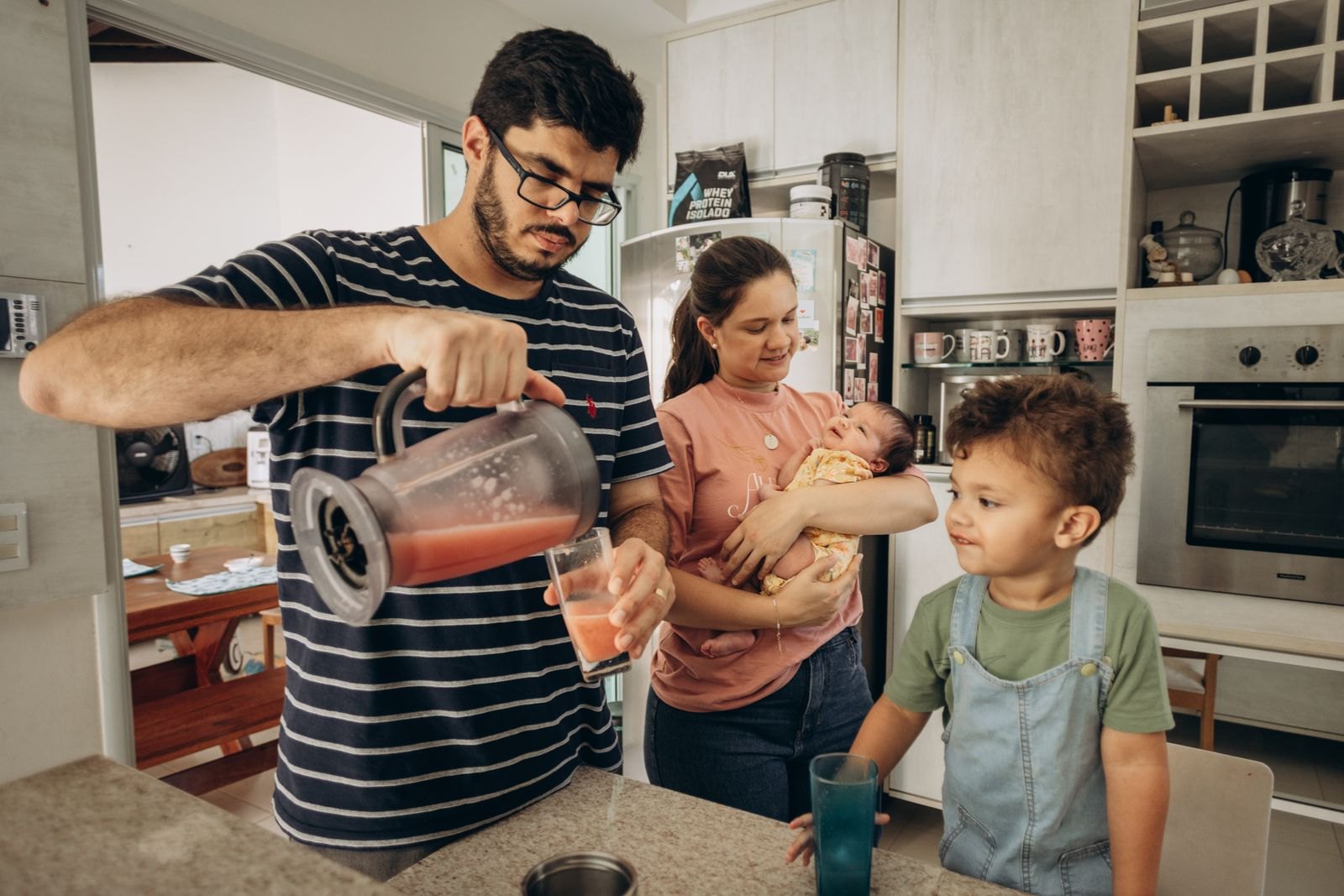Un padre prepara un zumo en la cocina mientras la madre sostiene al bebé recién nacido y el hermano mayor observa desde la encimera