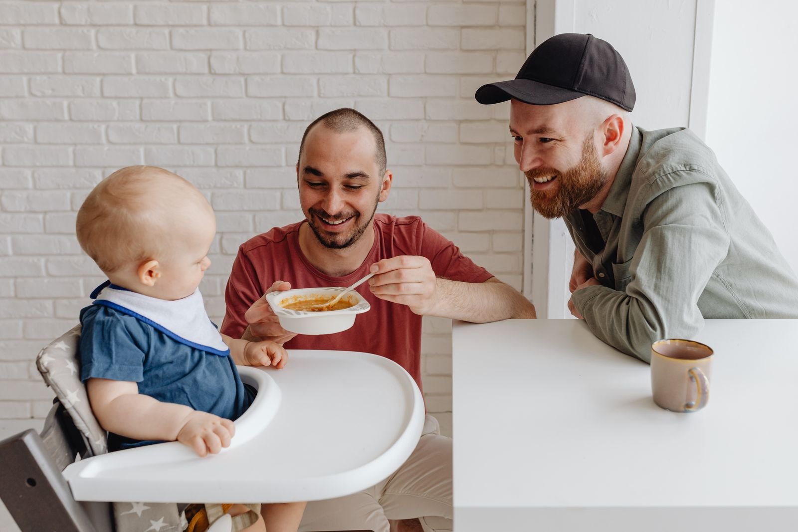 Un bebé en la trona siendo alimentado con cuchara por su padre, ambiente tranquilo y sonriente durante la comida familiar