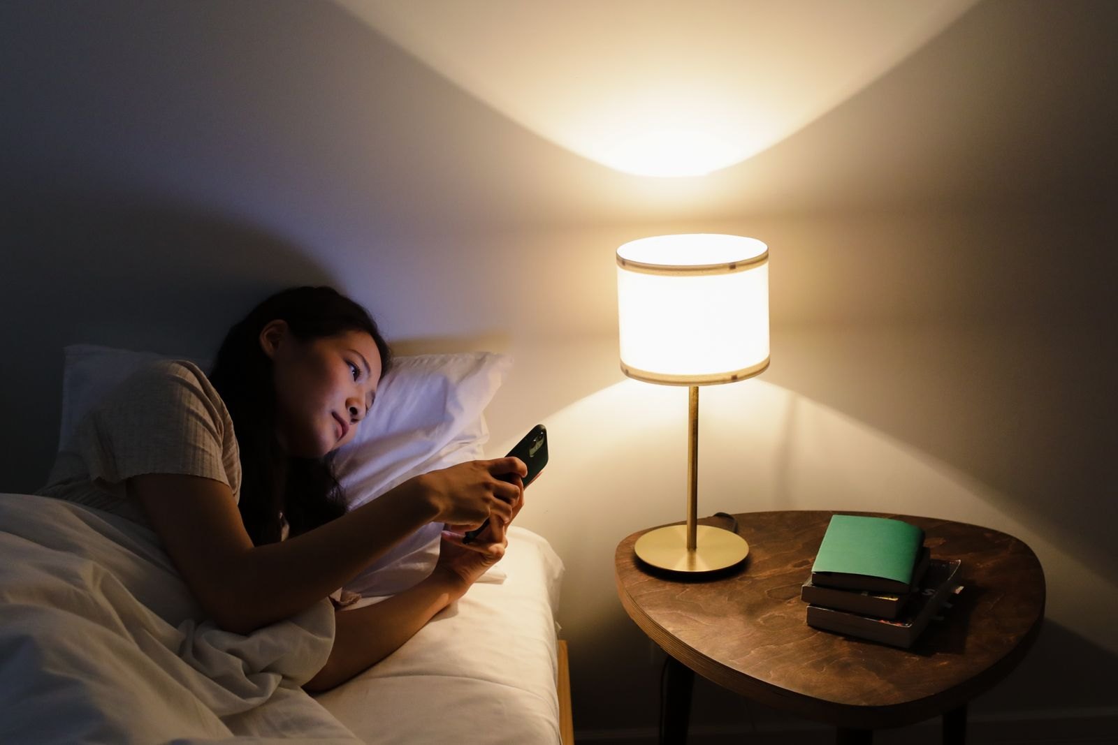 Una persona tumbada en la cama por la noche mirando la pantalla del móvil, con una lámpara de mesilla encendida de fondo