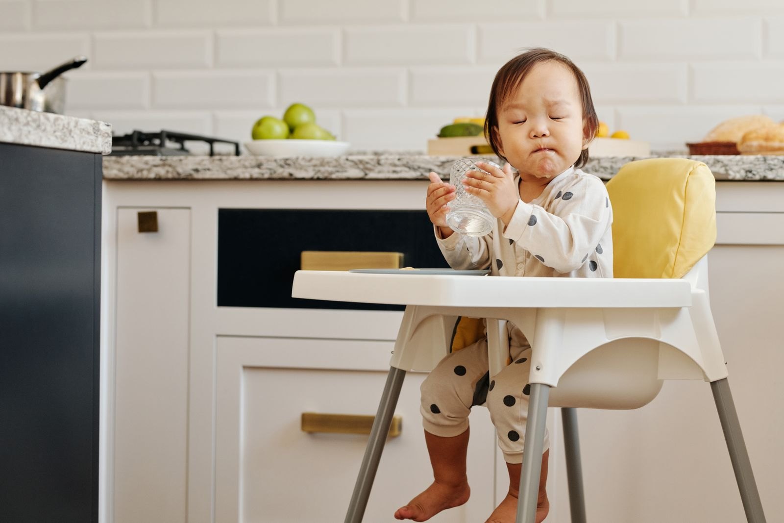 Bébé dans sa chaise haute fait la moue les yeux fermés en tenant son gobelet, une réaction fréquente face à un goût encore inhabituel