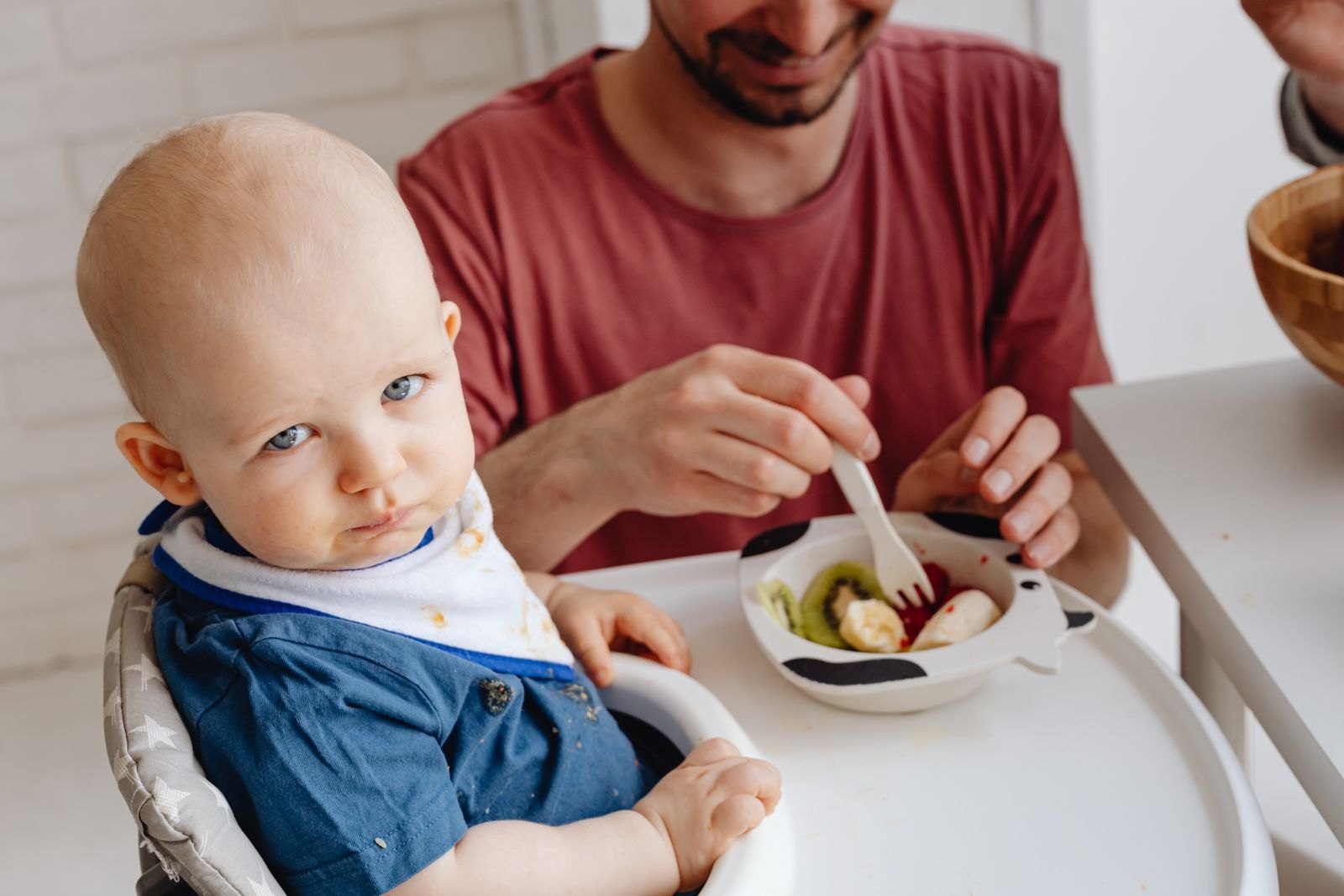 Bébé fait la moue dans sa chaise haute pendant qu'un parent lui tend une cuillère de fruits