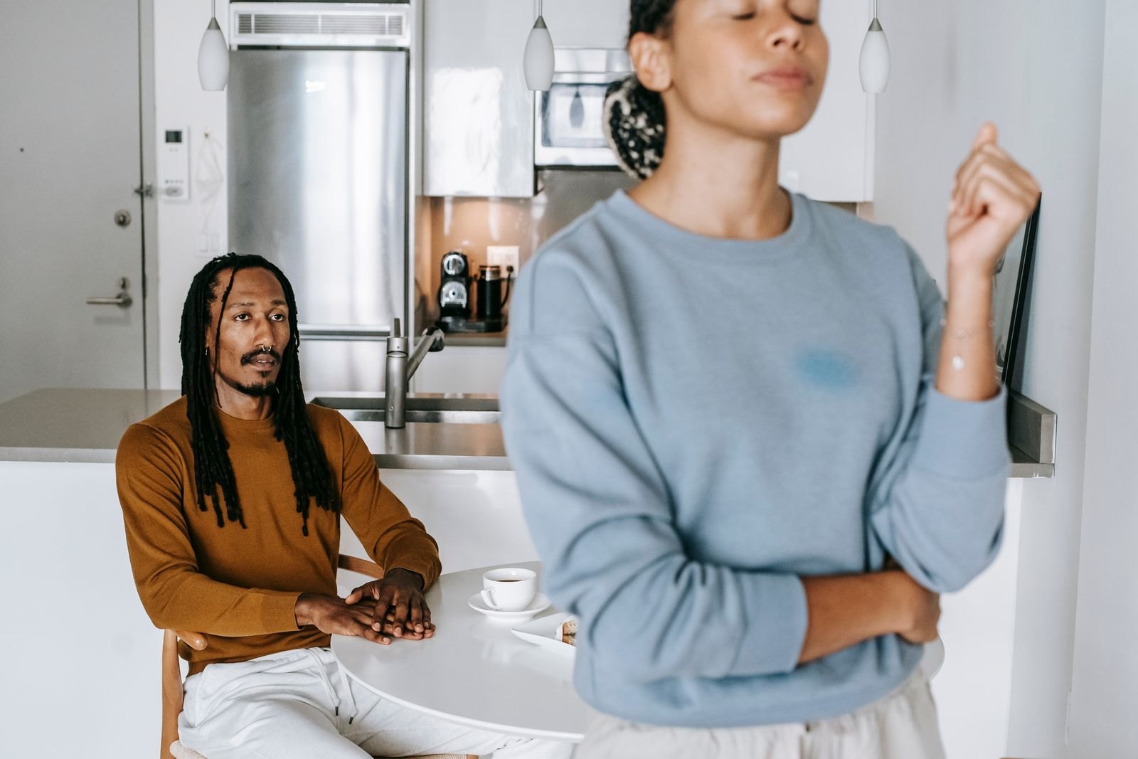 Un couple en tension dans une cuisine, l'un assis en silence, l'autre bras croisés en retrait