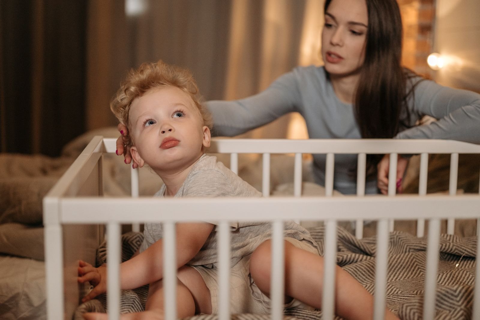Un enfant assis dans son lit se tourne vers sa mère qui pose la main sur son épaule, le soir