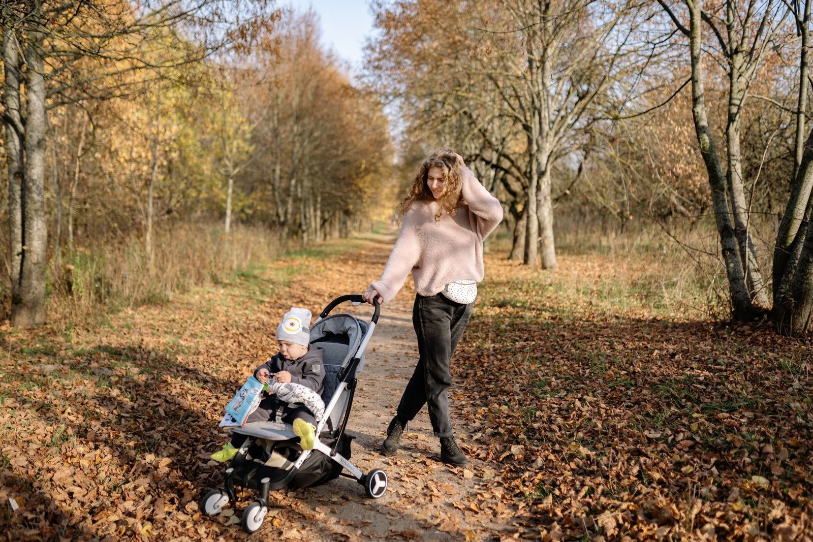 Une mère marche avec son jeune enfant installé dans une poussette, dans une allée bordée d'arbres en automne