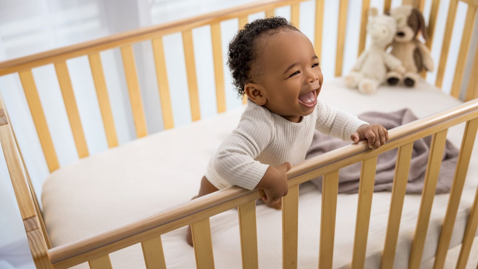 Bébé souriant debout dans son lit en bois, agrippé à la barrière, en train de s'entraîner à se tenir debout