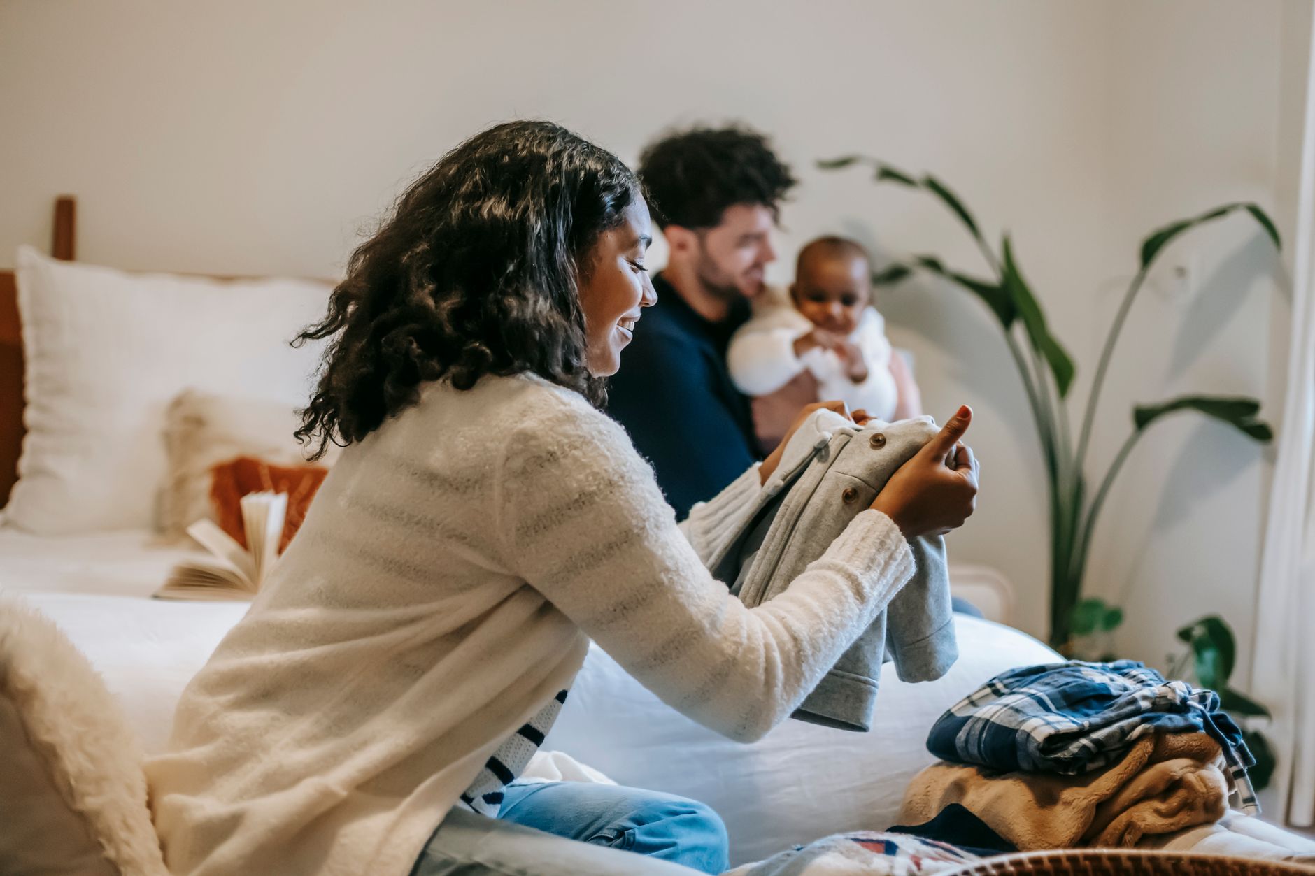 Une mère plie du linge de bébé au premier plan pendant qu'en arrière-plan, le père joue avec l'enfant sur le lit