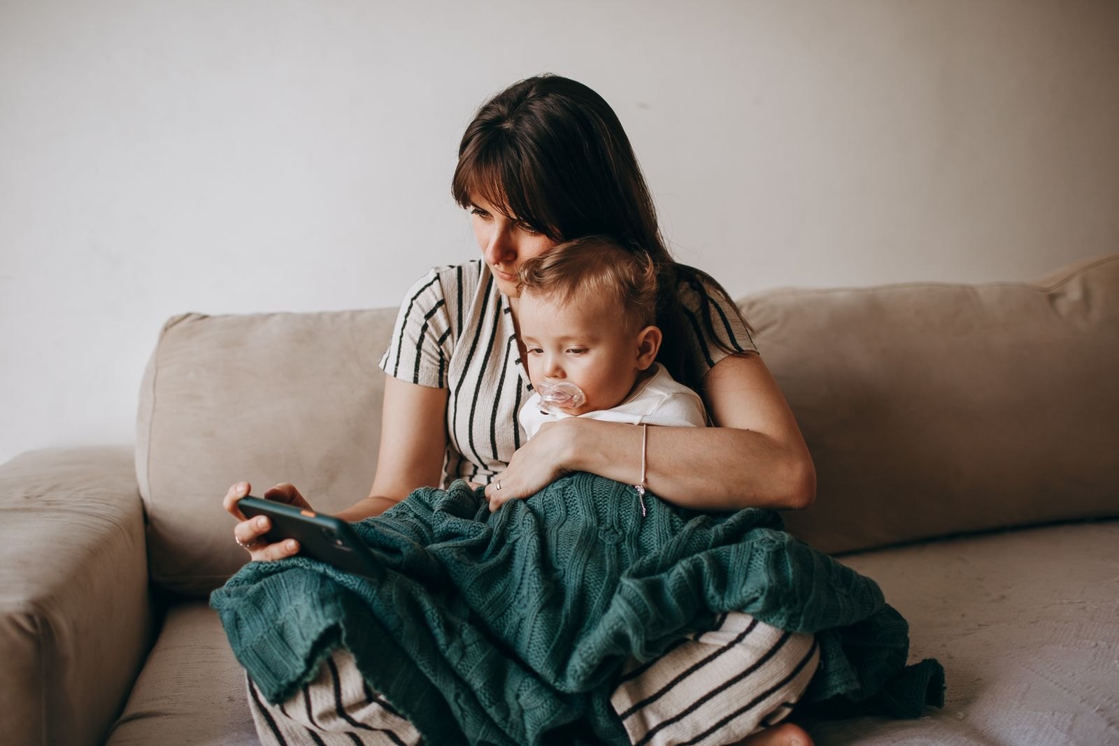 Une mère assise sur un canapé, tenant son bébé contre elle d'un air épuisé, le regard fixé sur son téléphone