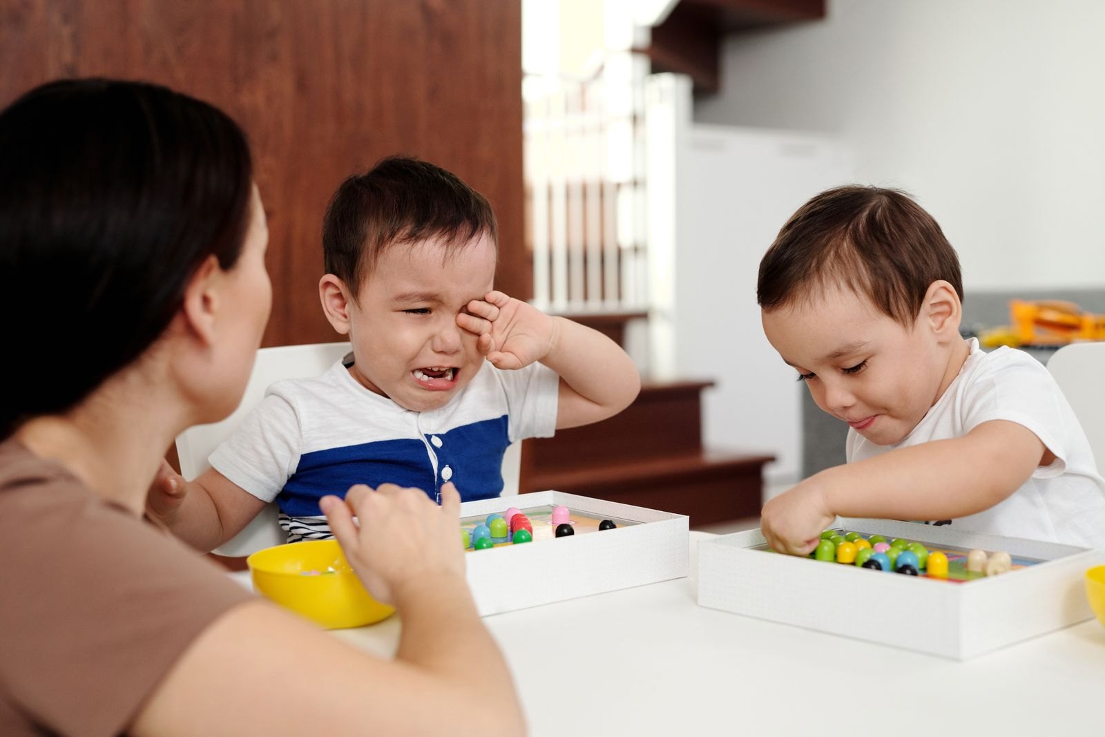 Deux tout-petits assis à une table avec un adulte, l'un en pleurs et l'autre concentré sur son jeu
