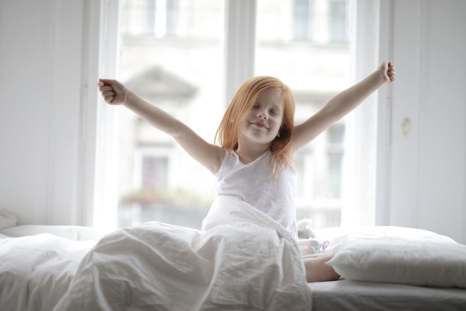 Una bambina si stiracchia felice al risveglio davanti a una finestra luminosa