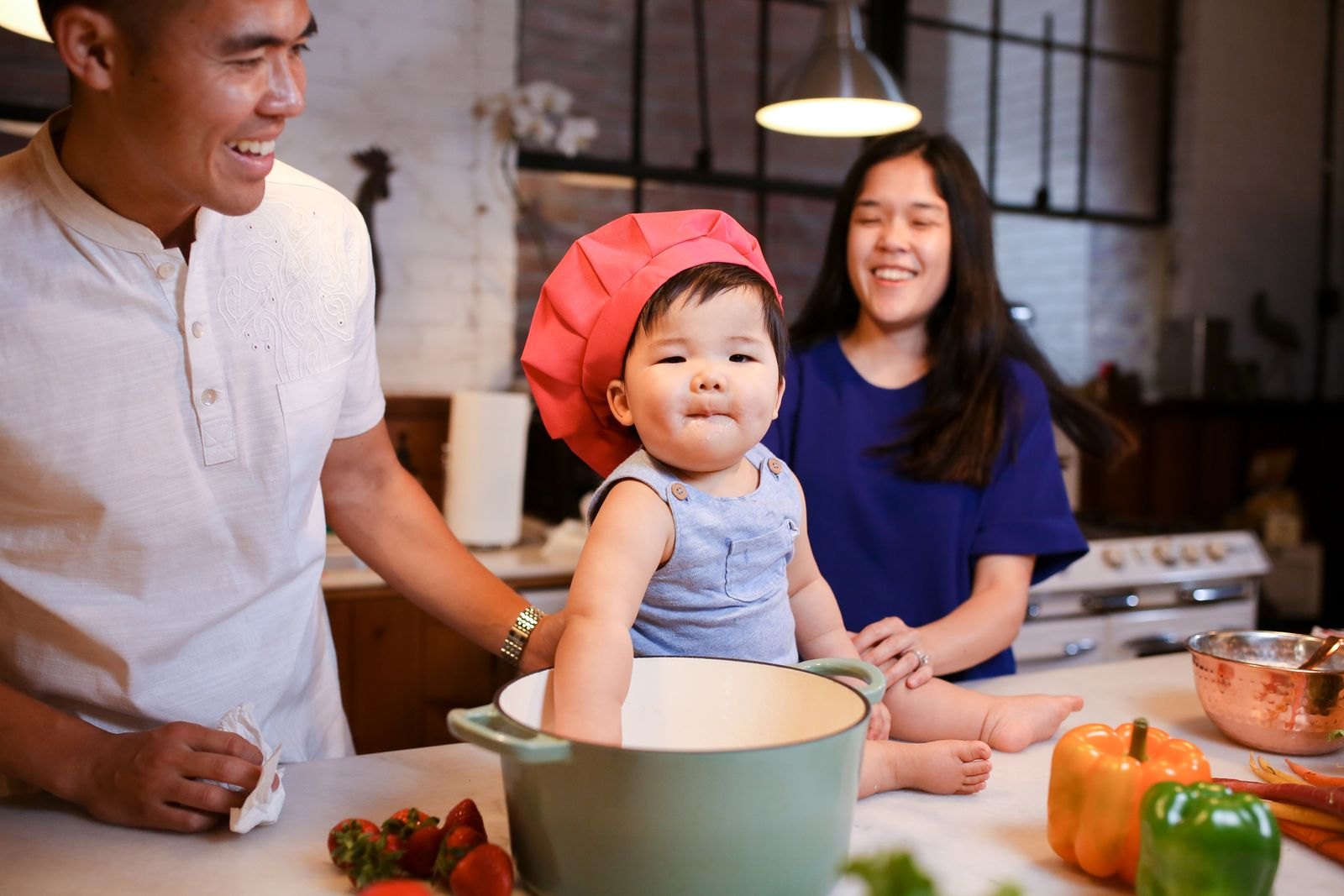 Bambino con cappello da chef seduto su un piano cucina circondato da verdure fresche, genitori sorridenti accanto