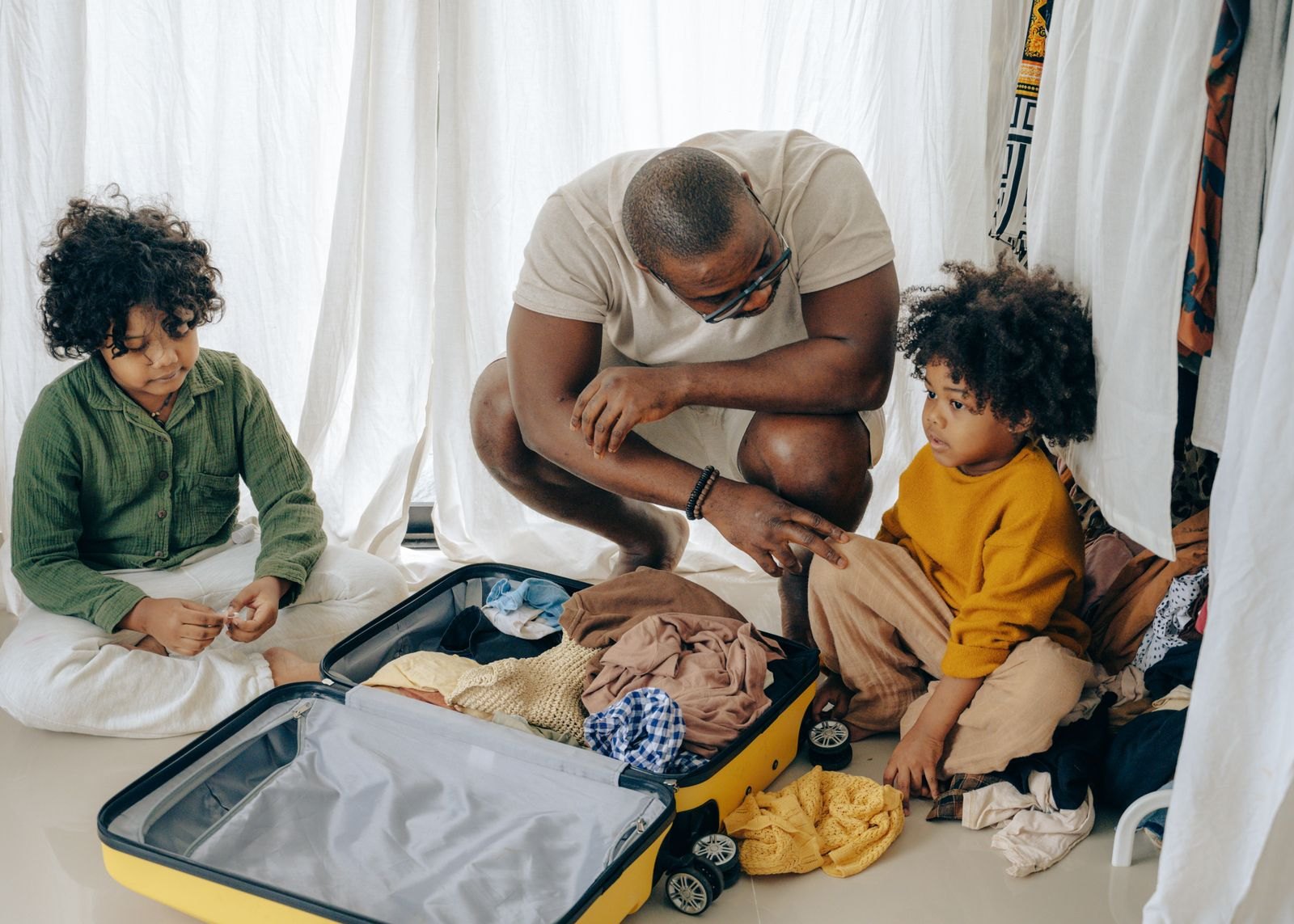Un padre prepara la valigia mentre due bambini lo osservano e giocano accanto, coinvolti nella partenza