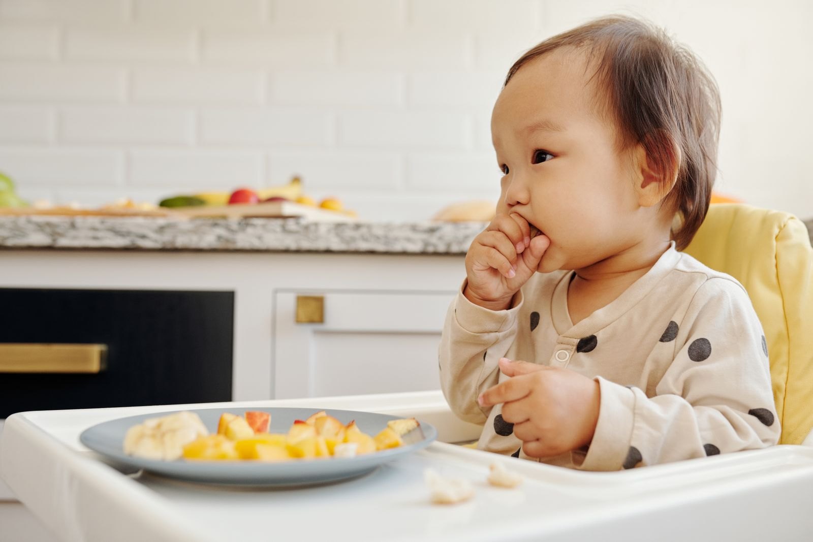 Un bambino seduto nel seggiolone mangia pezzi di frutta con le mani, davanti a un piatto con banana e pesca