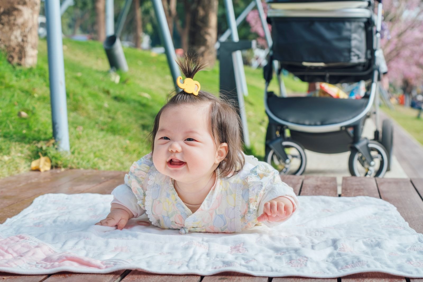 잔디밭 옆 담요 위에 엎드려 웃는 아기, 뒤에 접힌 유모차