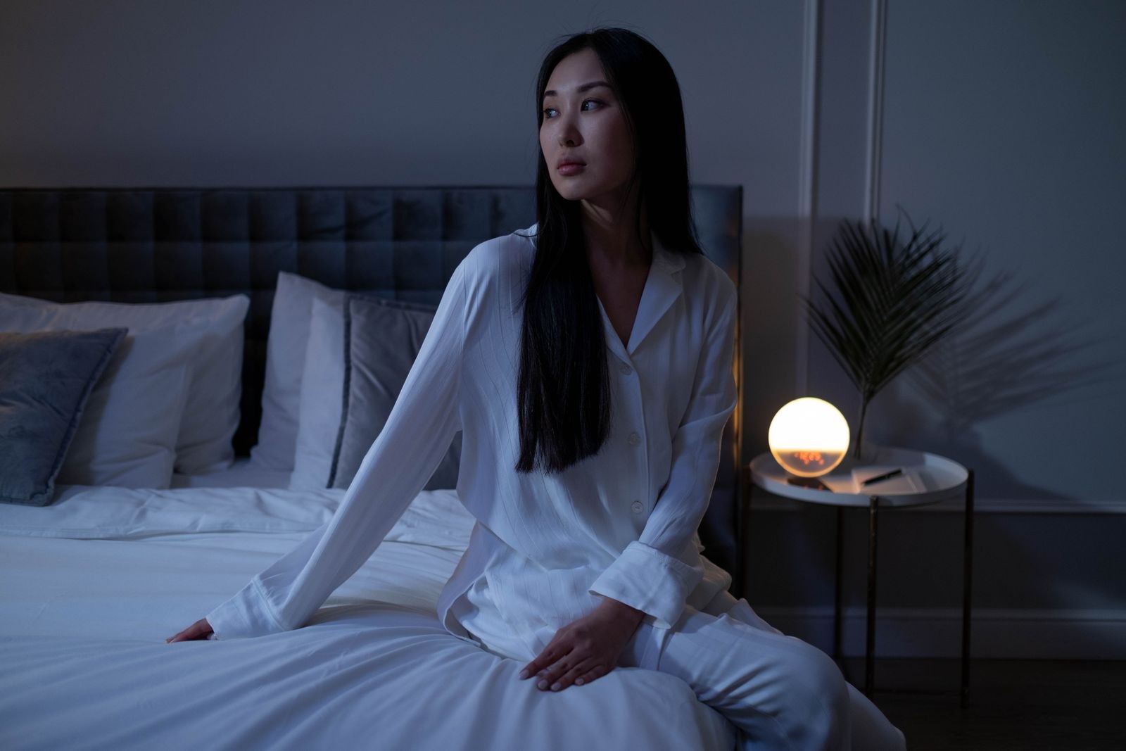 어두운 침실 침대에 앉아 있는 잠옷 차림의 여성과 옆의 무드등