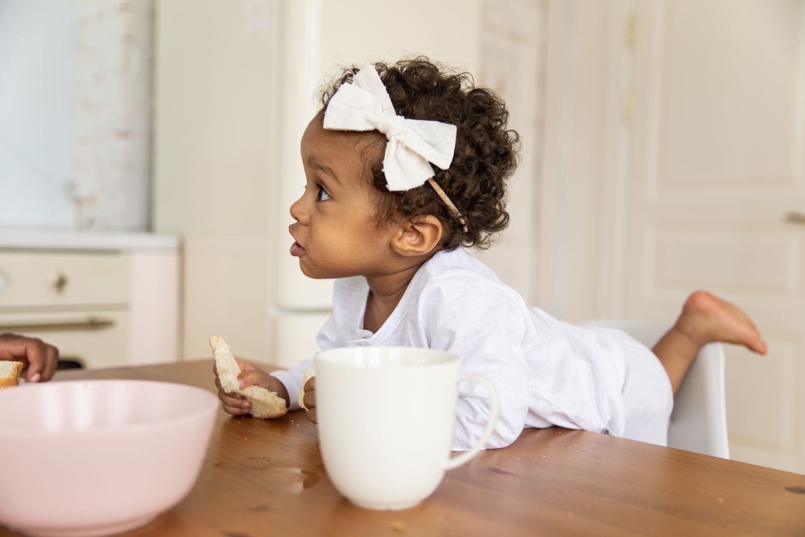 Baby in de kinderstoel pakt zelf stukjes eten van het blad terwijl de ouders op de achtergrond in de keuken bezig zijn