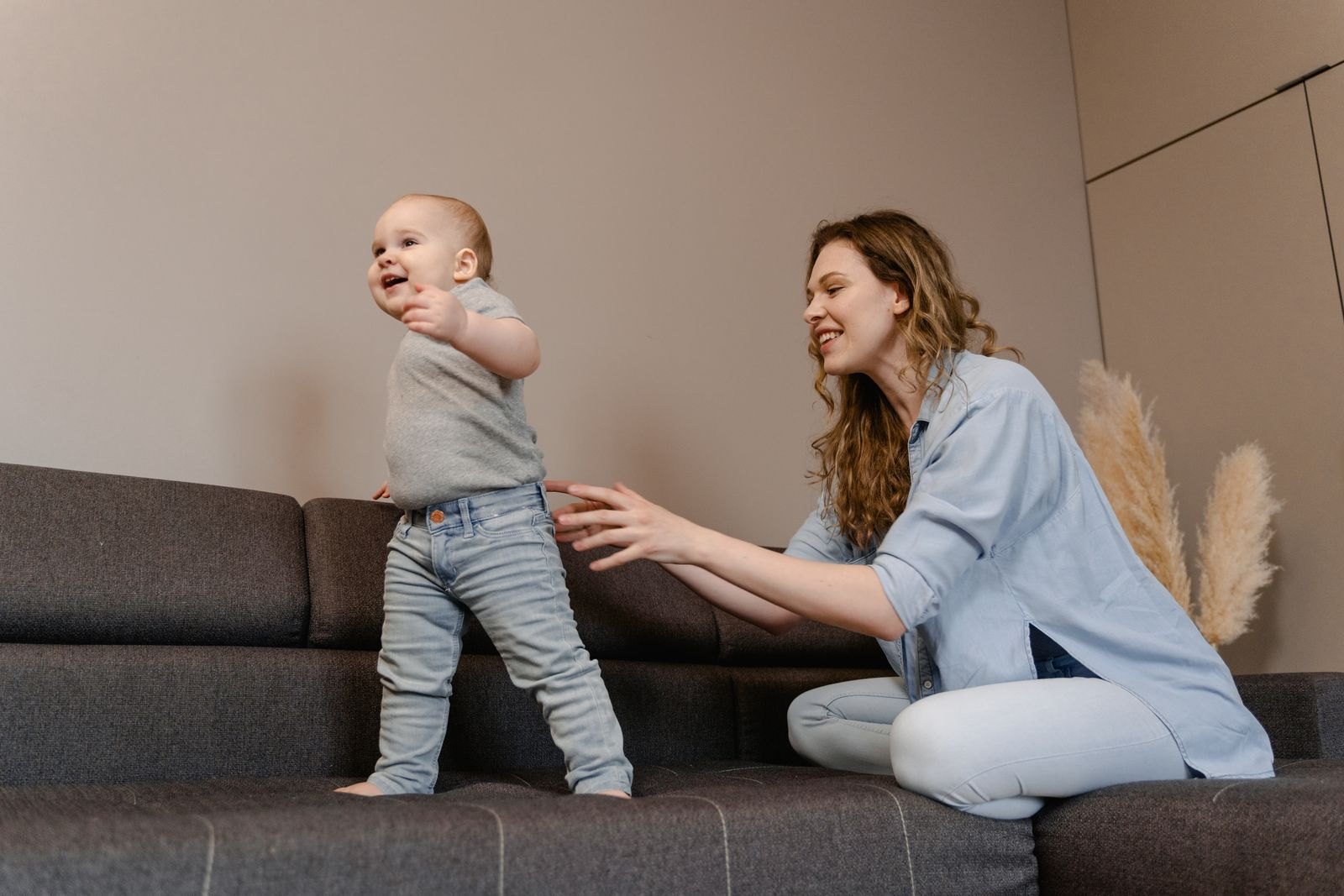 Een baby oefent met lopen langs de bank terwijl een ouder toekijkt en de handjes vasthoudt