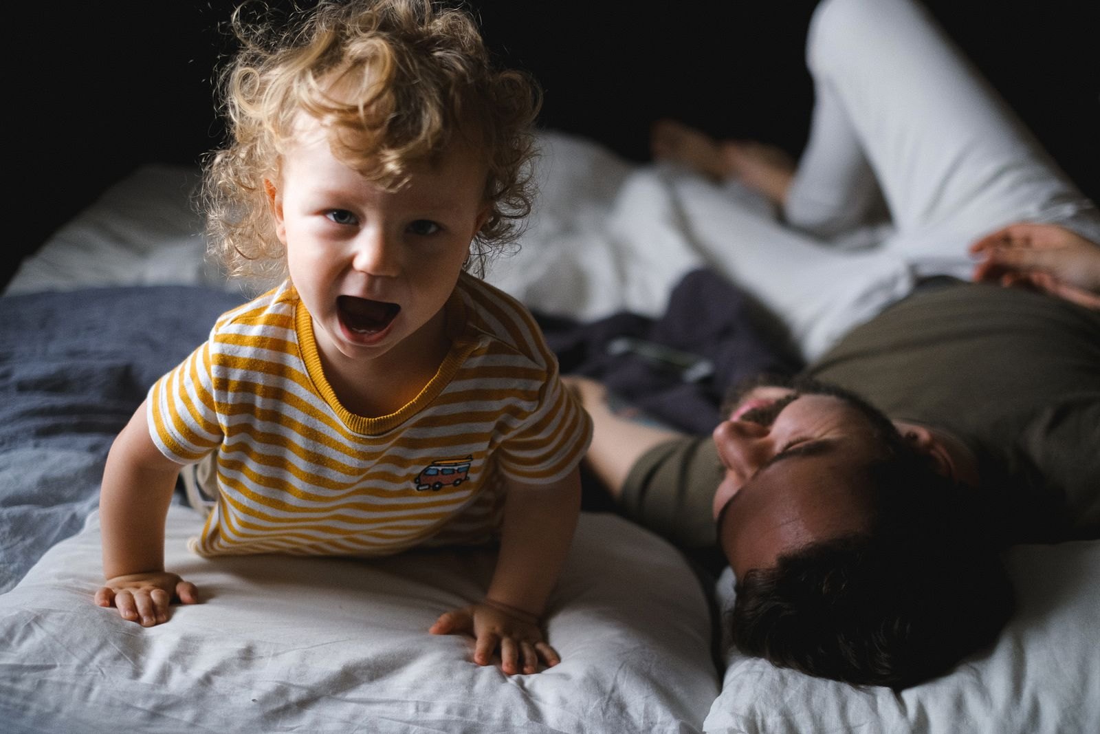 Een peuter maakt klaarwakker geluid op bed terwijl een uitgeputte vader er met gesloten ogen naast ligt
