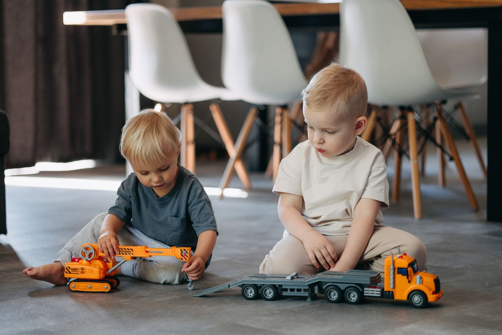 Twee jonge kinderen zitten naast elkaar op de grond, elk verdiept in hun eigen speelgoedvoertuig
