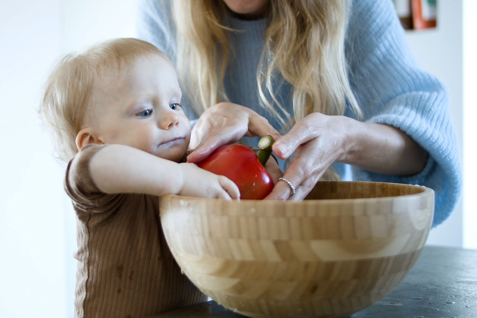 En liten bebis utforskar en röd paprika med händerna i en skål medan en förälder håller i grönsaken