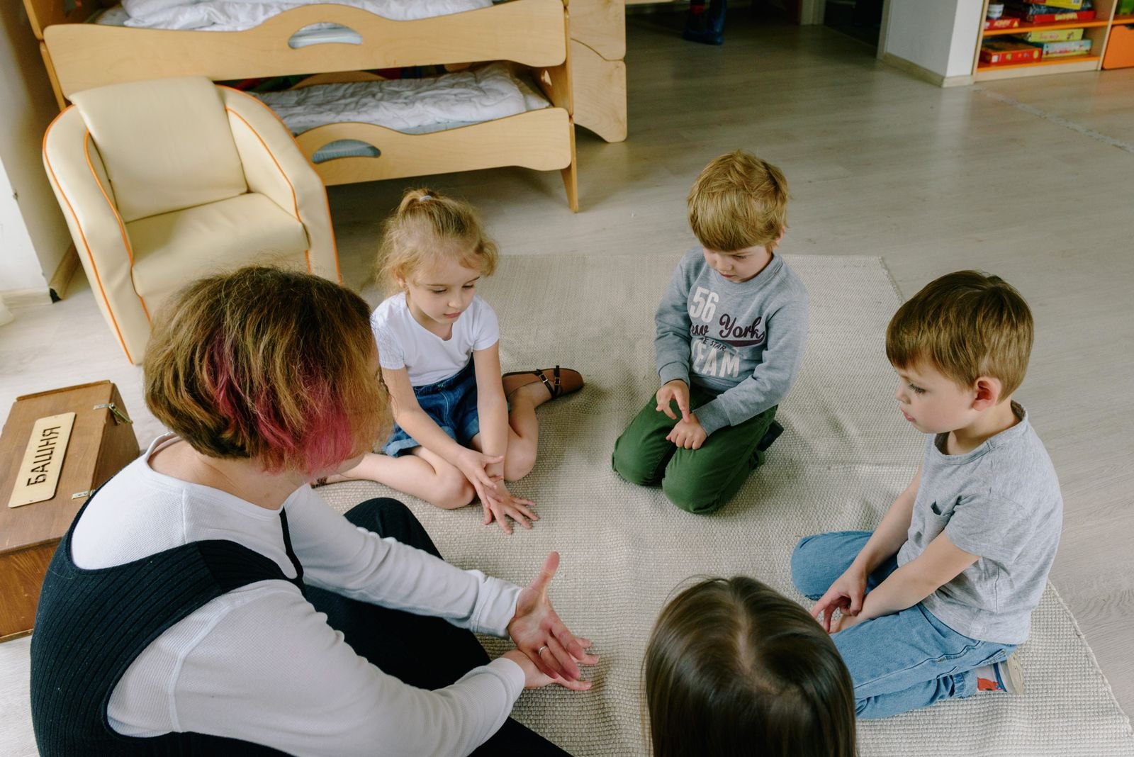 Barn sitter tillsammans med en pedagog i ett rum med sovplatser i bakgrunden, en del av förskolans vardagliga rutin