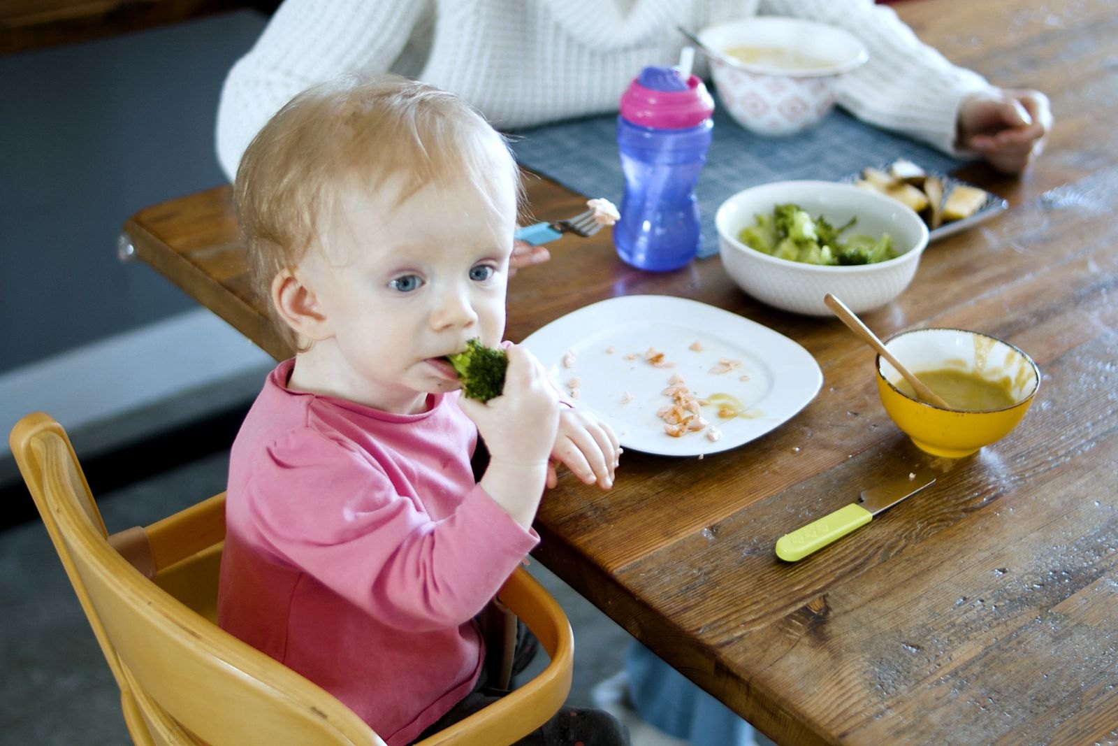 Ett barn äter broccoli med fingrarna vid familjens matbord, med både puré och hela bitar på bordet