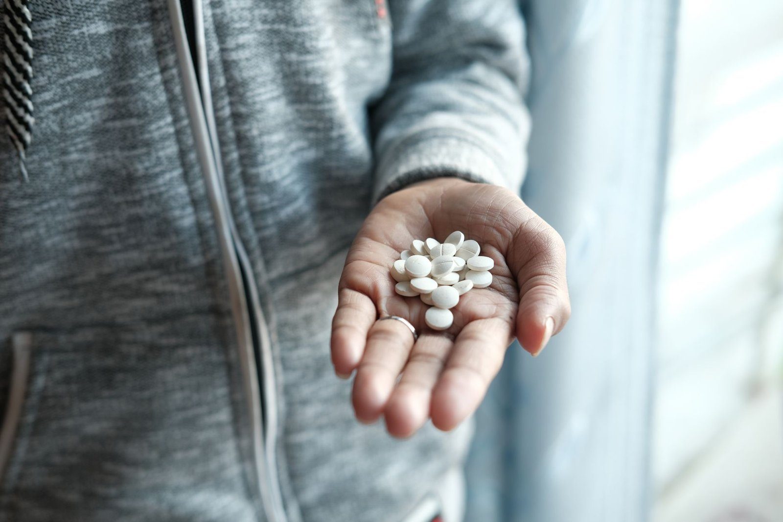 An open palm holding a small heap of white oval tablets, the person wearing a grey zip-up jacket