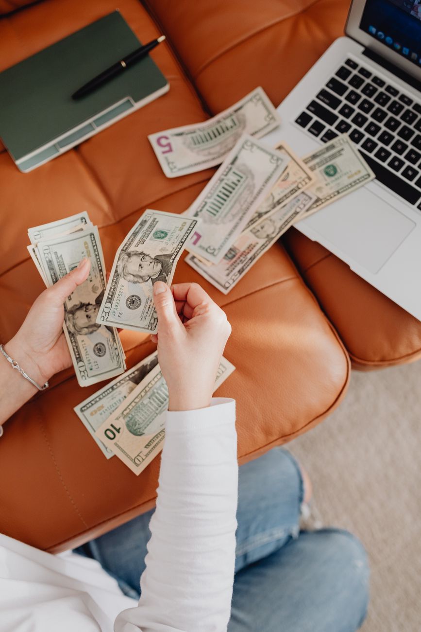 Person counting cash bills of varying denominations next to an open laptop, representing tracking variable self-employed or commission-based income