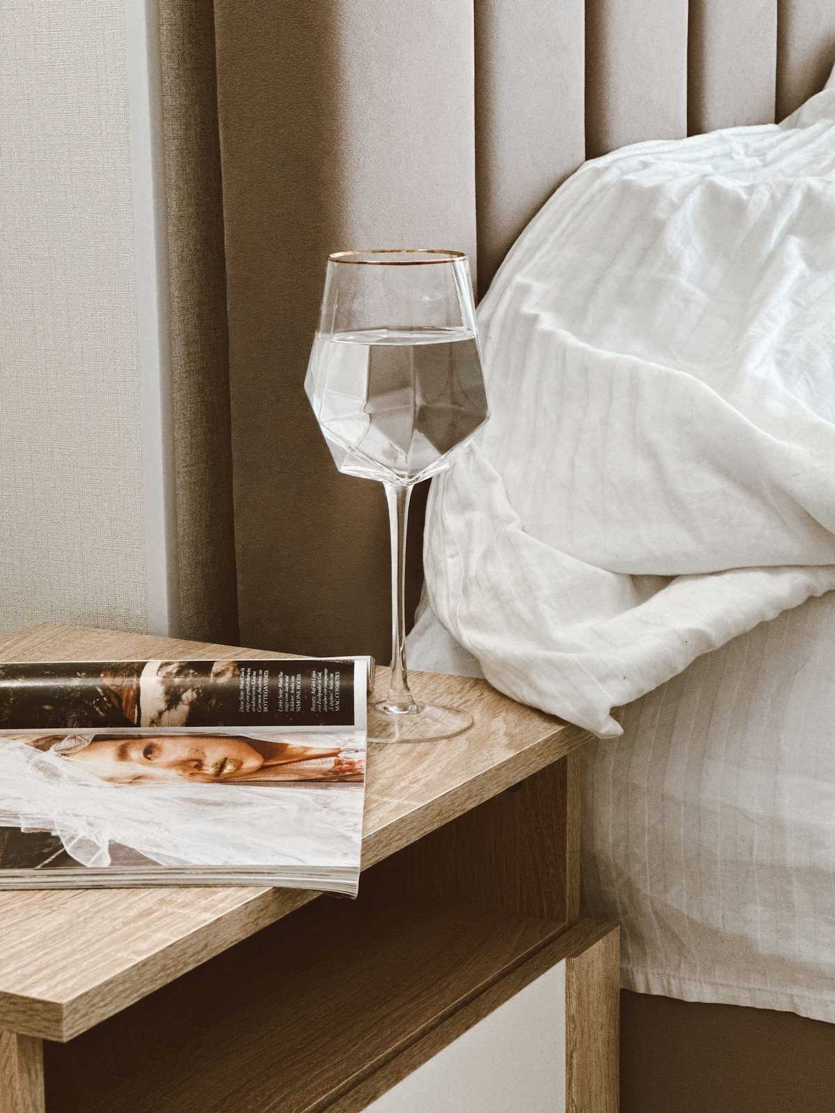 A faceted glass goblet half full of water on a light oak bedside table beside an open magazine, with a rumpled white duvet behind