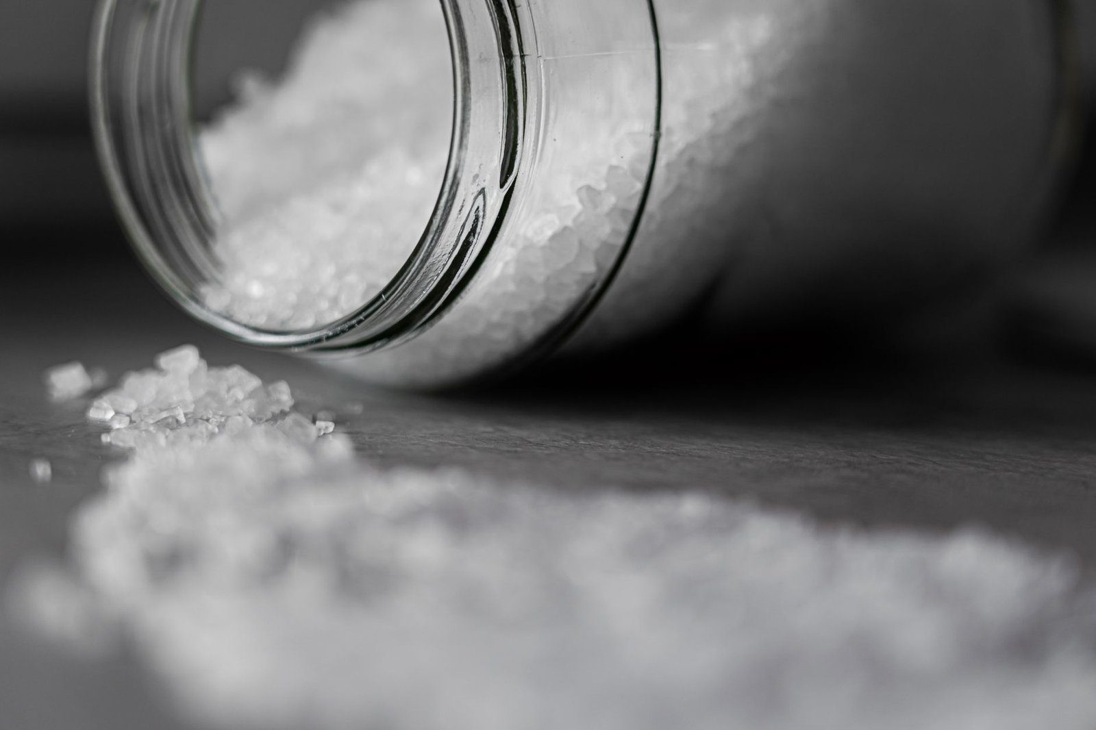 A glass jar tipped on its side with coarse salt spilling into a pile on a dark stone surface, in black and white