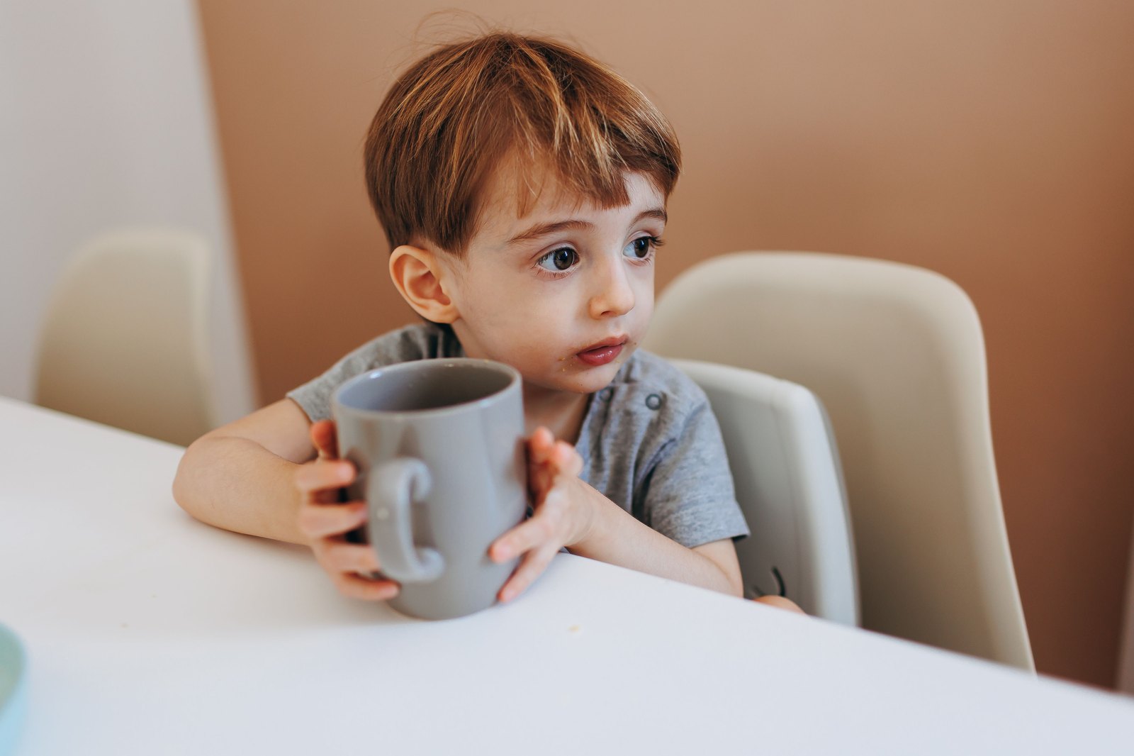 Un tout-petit tient une tasse à deux mains à table et boit seul
