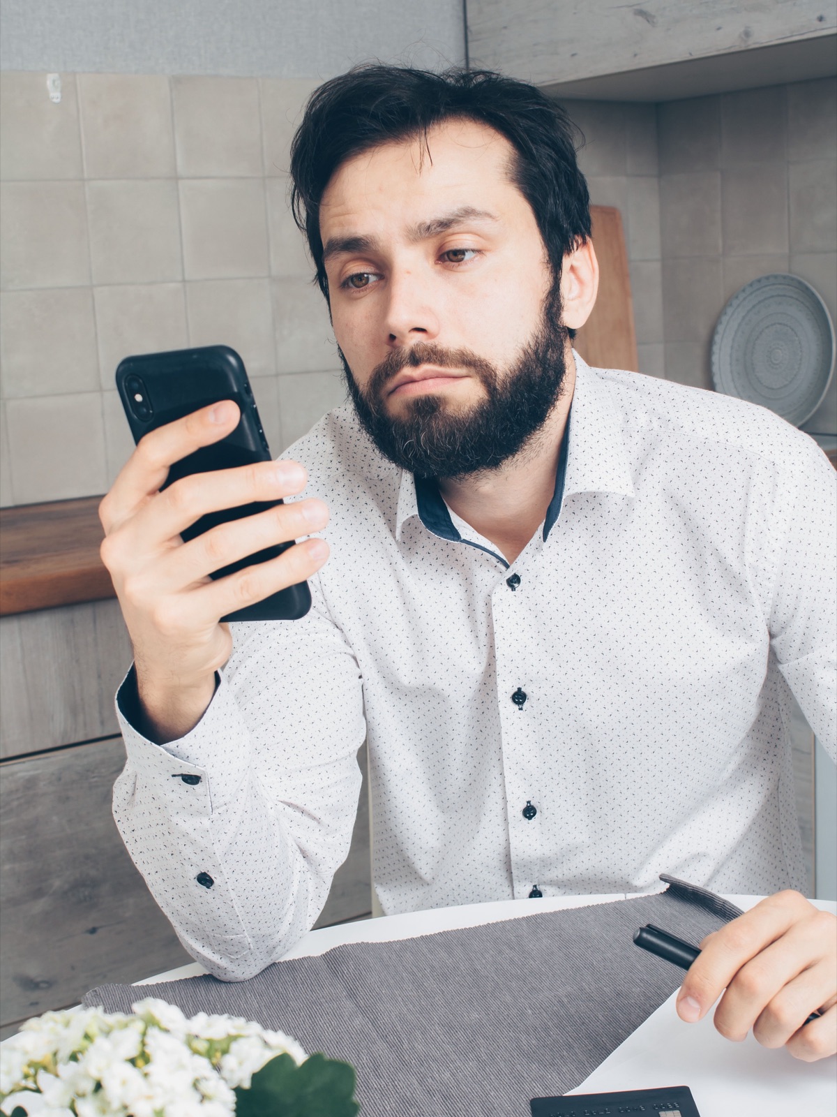 男性がキッチンのテーブルでクレジットカードと書類を前に、真剣な表情でスマートフォンの画面を見ている