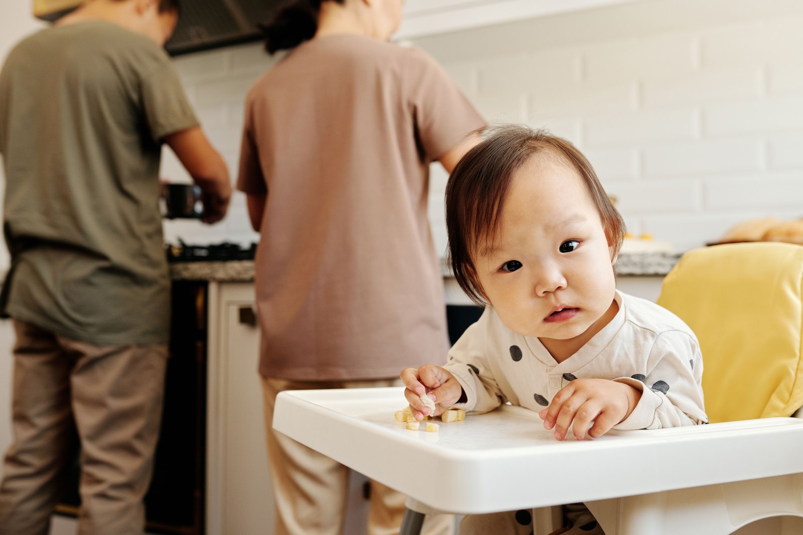 ハイチェアでカメラを見つめる赤ちゃん、背後で両親が調理している