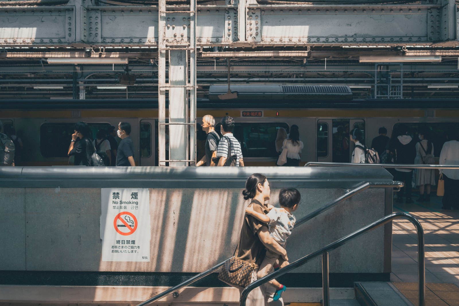 駅の階段で子どもを抱えながらトートバッグを肩にかけて上る母親