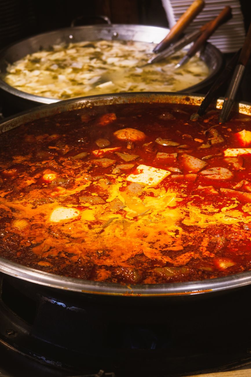 가스불 위 큰 냄비에 두부와 고기가 든 붉은 김치찌개가 끓고 있고 뒤쪽에 다른 국 냄비가 놓인 사진