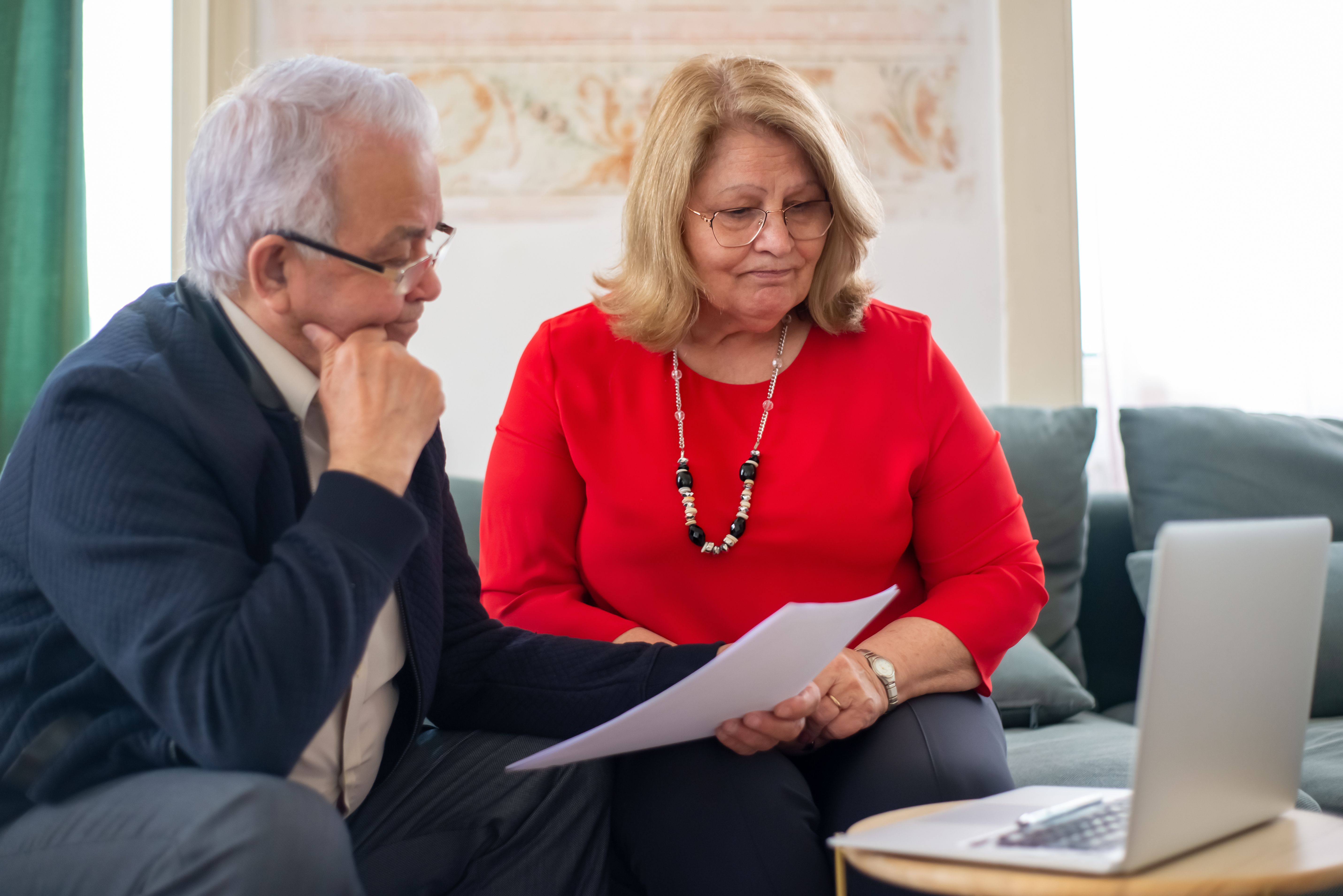 노년의 부부가 서로 기대어 앉아 테이블 위 서류를 앞에 두고 카메라를 보며 미소짓고 있다